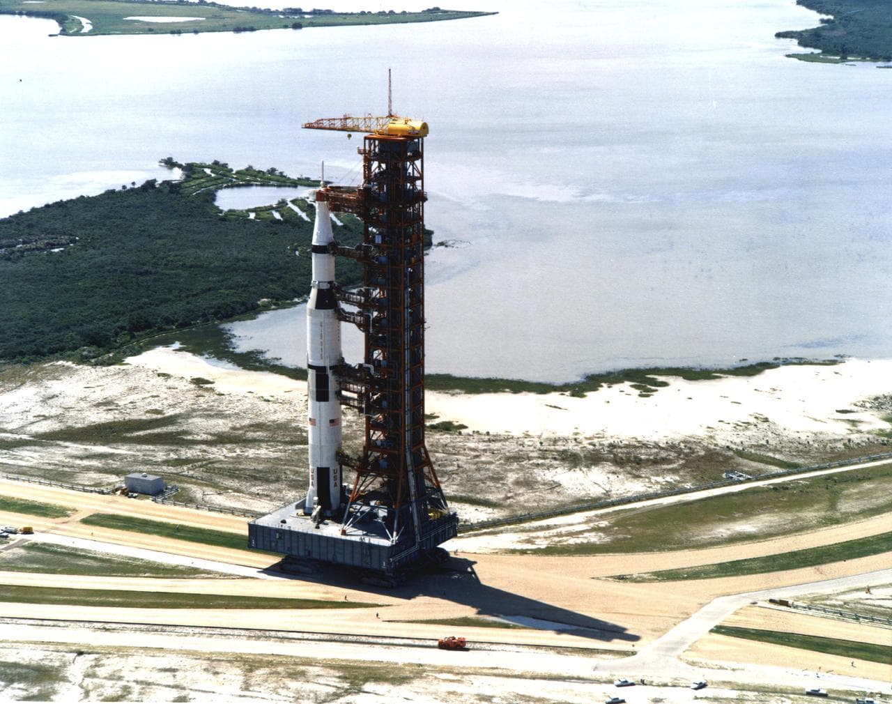 An aerial view of the Saturn V rocket and its launch tower riding the crawler-transporter along the crawlerway toward the launch pad.