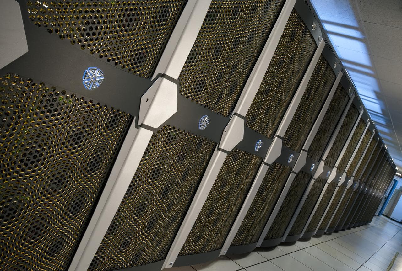 A long aisle between rows of supercomputer cabinets, cables and status lights running away toward the far wall.