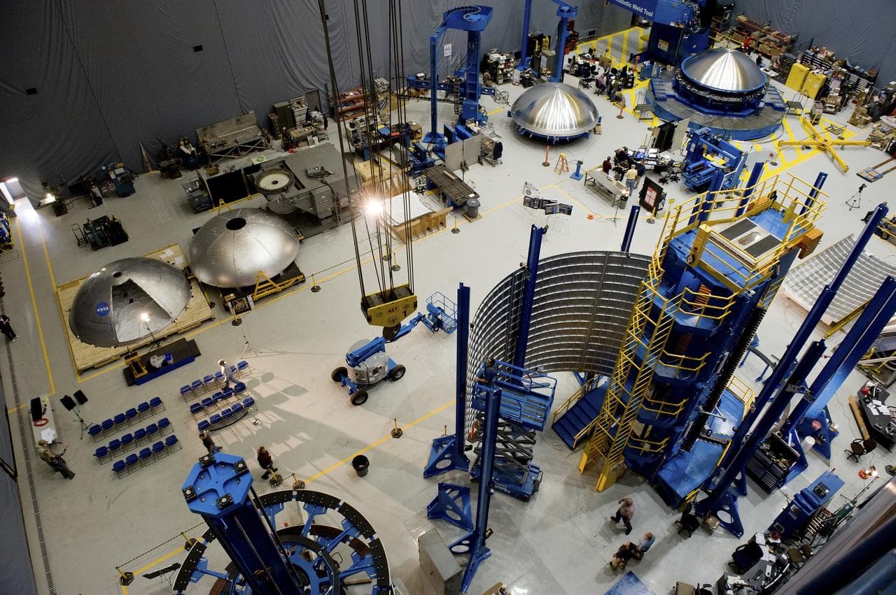 Overhead view of a factory floor where large spun metal tank domes and a curved panel stand among blue welding tooling.