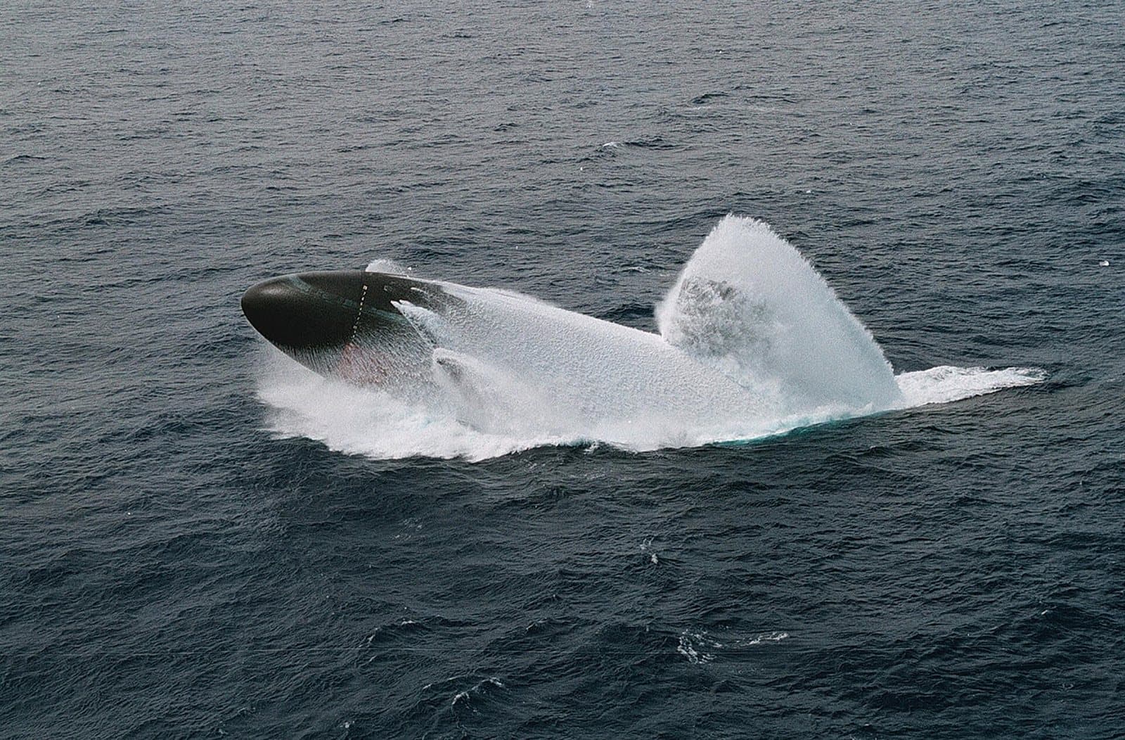 A submarine bursting bow-first out of the open ocean during a surfacing drill, throwing up a wall of white water.