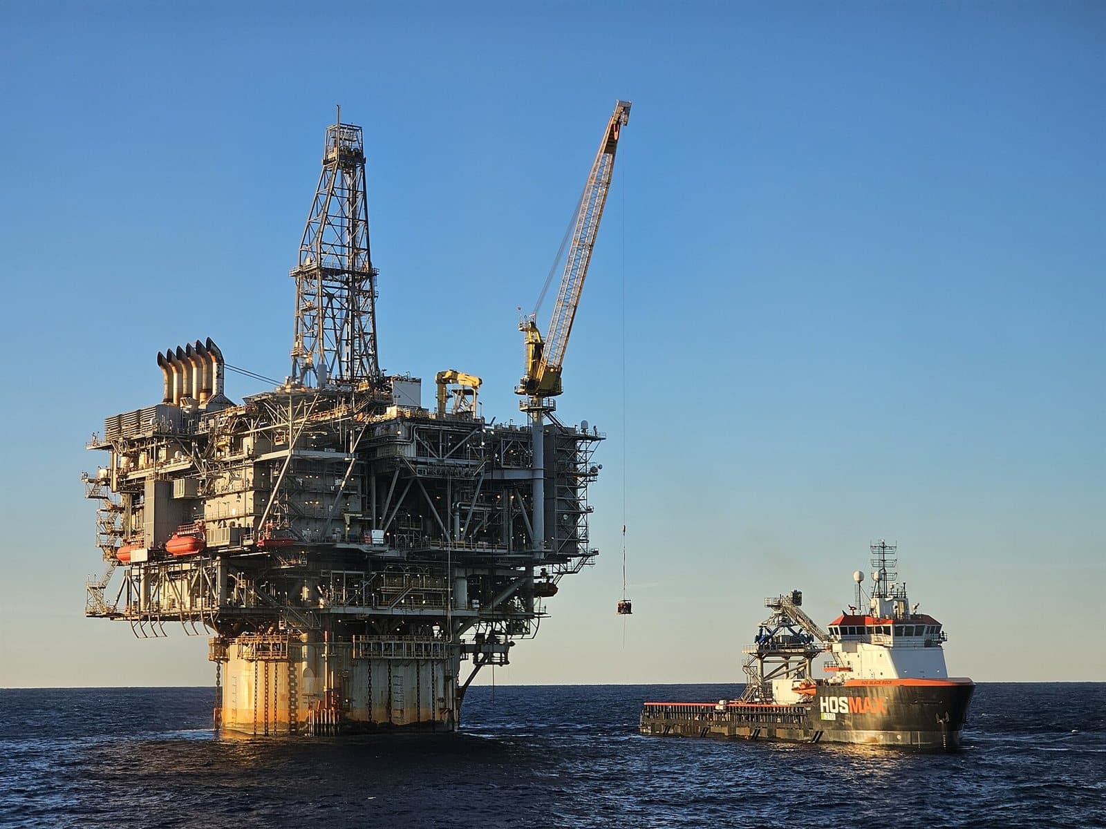 An offshore oil platform standing on its legs above the sea, a crane swinging a personnel basket across open water.