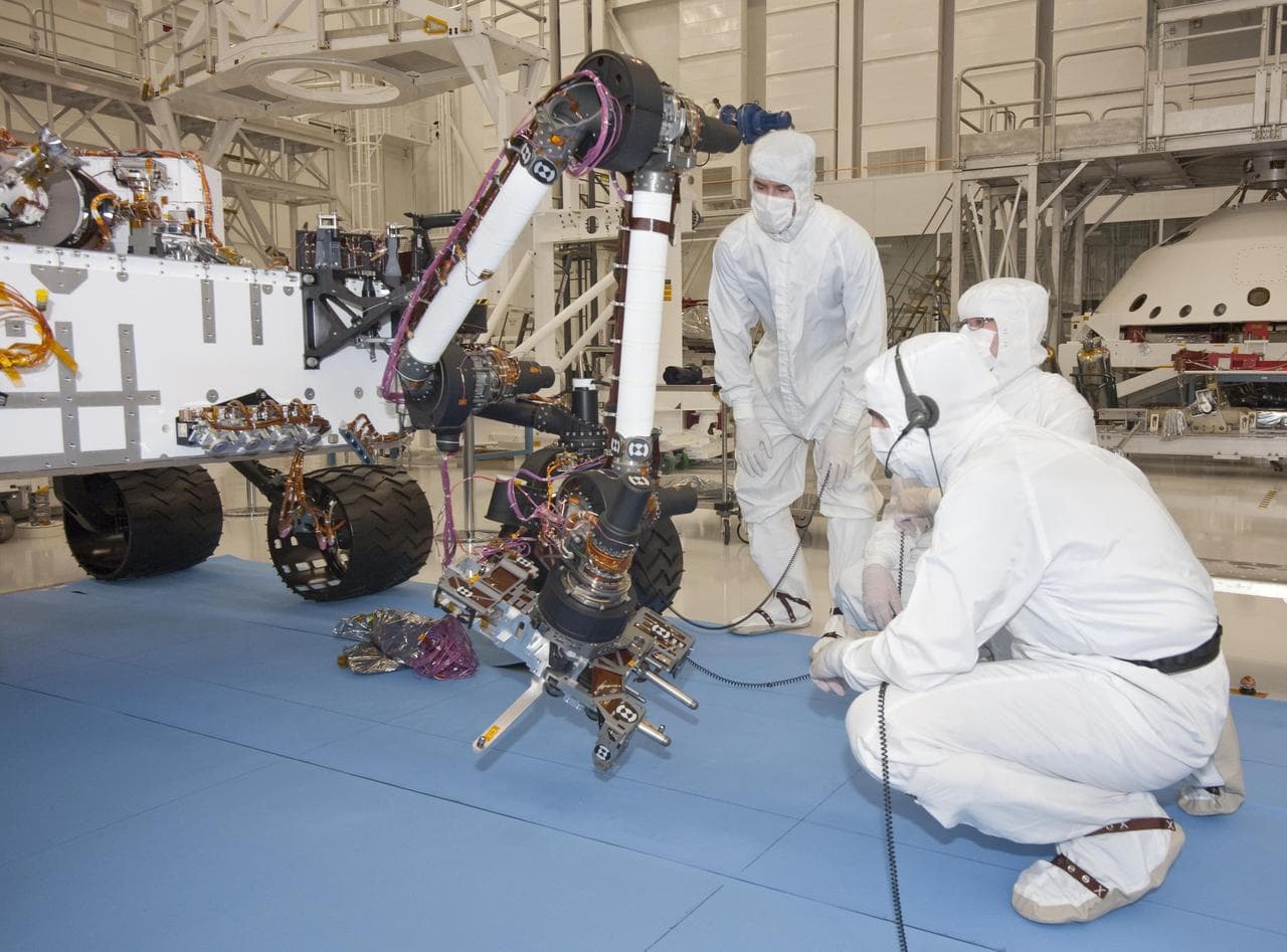 Engineers in cleanroom suits working around the Curiosity rover as it extends its robotic arm.