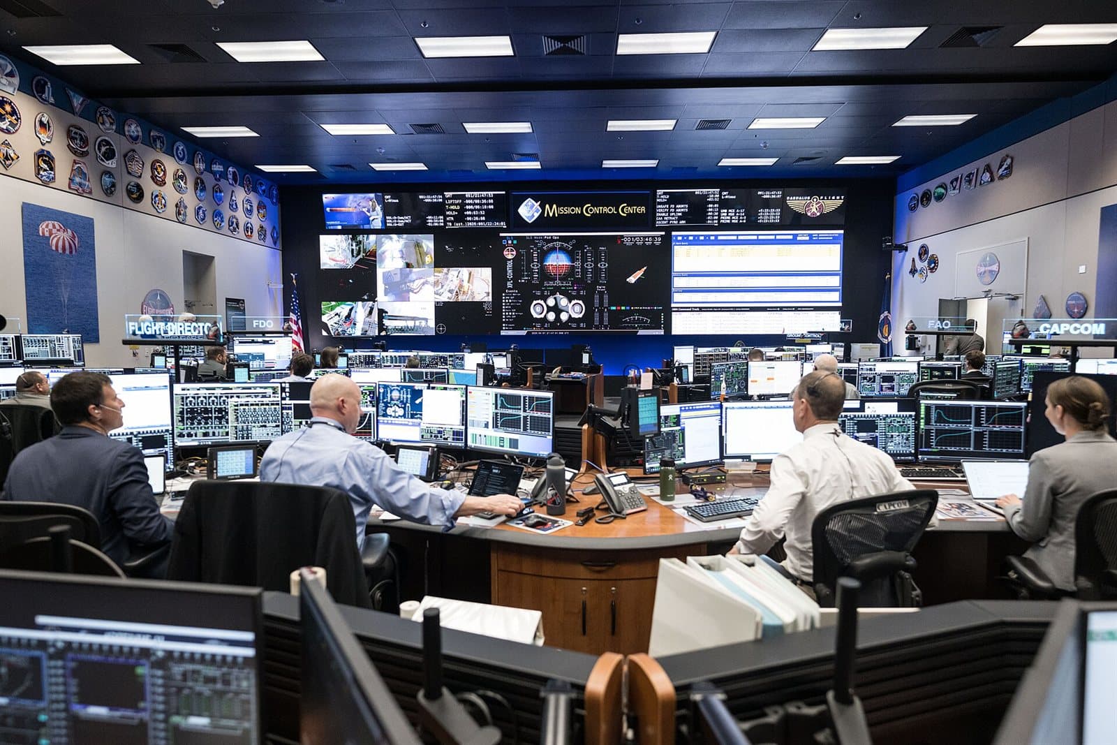Flight controllers at consoles in a mission control room, each position banked with screens under a wall of larger displays.