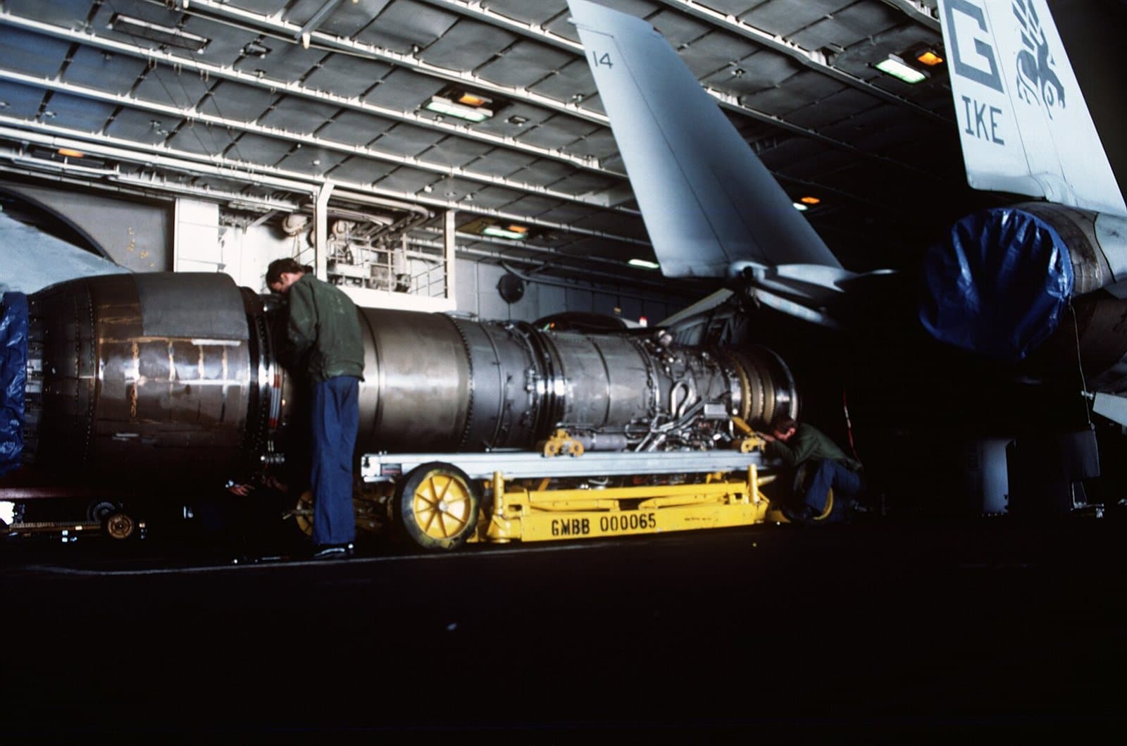 Ground crew guiding a large jet engine into the open engine bay of a fighter aircraft in a hangar.